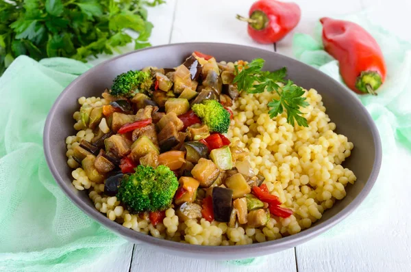 Ensalada vegana caliente con cuscús y verduras fritas (berenjena, calabacín, cebolla, pimiento dulce, brócoli) en un tazón de cerámica sobre un fondo de madera blanca. Nutrición adecuada . — Foto de Stock