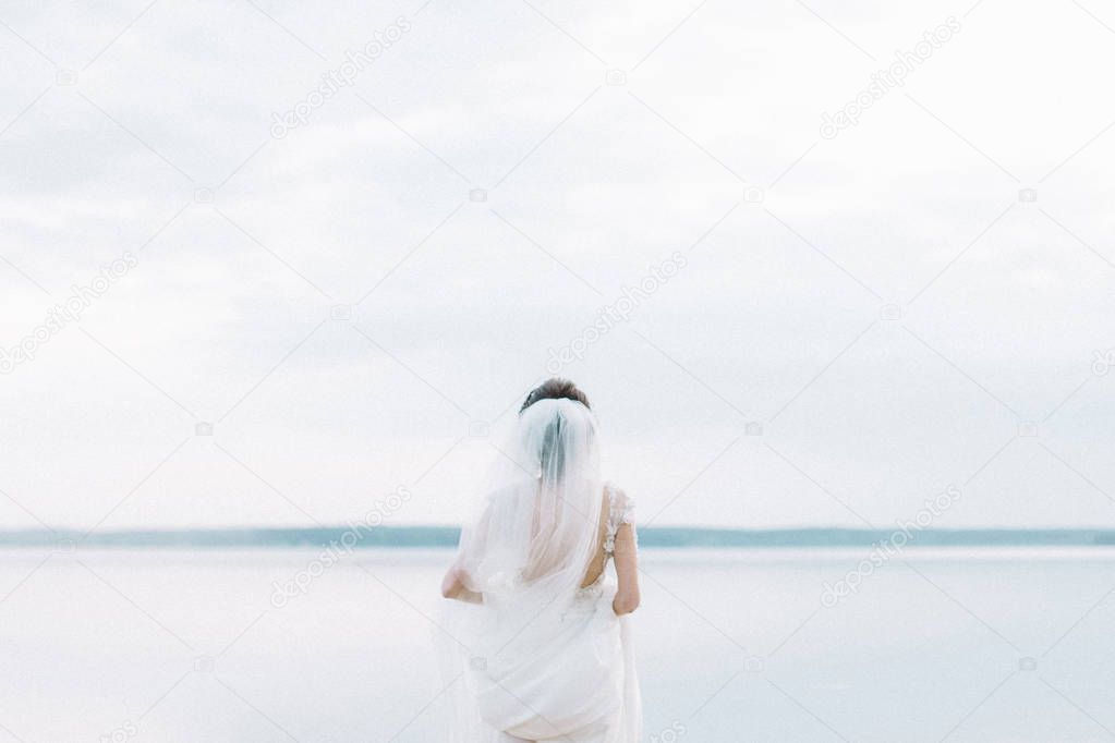 Back view on bride in wedding dress look at horizon near lake. Minimal bridal fashion background.