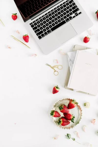 Female home office desk workspace with laptop, notebook, lipstick, fresh raw strawberries and rose flower buds on white background. Flat lay, top view.