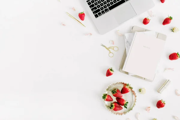 Home office desk workspace with laptop, notebook, lipstick, fresh raw strawberries on white background. Flat lay, top view.