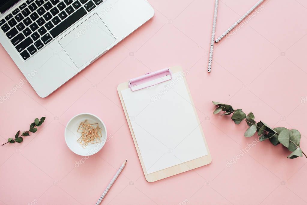 Flat lay office workspace with blank laptop, clipboard, eucalyptus branches on pink background. Top view minimal mock up template concept.