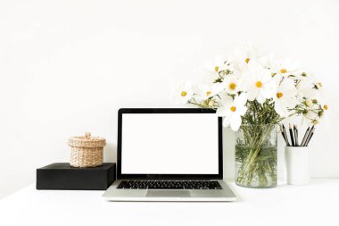 İş, serbest çalışma, blog yazarı konsepti. Model yap, boş, kopyala alanı. Diz üstü bilgisayar beyaz masanın üzerinde vazoda papatyalar, siyah kutu, saman sepet ile beyaz duvara karşı duruyor.. 