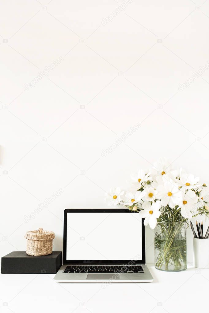 Business, freelance, blogger concept. Mock up, empty, copy space. Laptop standing on white table against white wall with daisies in vase, black box, straw basket. 
