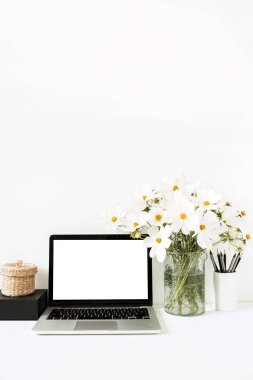 İş, serbest çalışma, blog yazarı konsepti. Model yap, boş, kopyala alanı. Diz üstü bilgisayar beyaz masanın üzerinde vazoda papatyalar, siyah kutu, saman sepet ile beyaz duvara karşı duruyor.. 