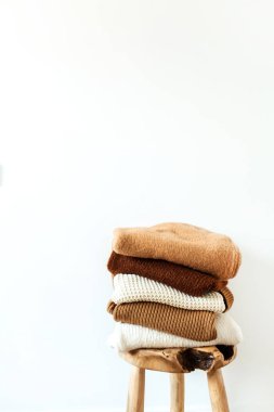 Kadın moda kompozisyonu. Sıcak kış dişisi yün örgü süveterler, kazaklar beyaz arka planda tahta tabureye istiflenmiş. Dergi, blog, sosyal medya için modern kıyafet konsepti.