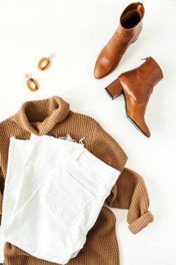 Kadın kıyafetleri, beyaz arka planda aksesuarlar. Düz, üst görünüm en az moda yaşam tarzı kolajı. Kahverengi balıkçı yaka kazak, ayakkabı, kot pantolon, küpe. Moda blogcusu kavramı.