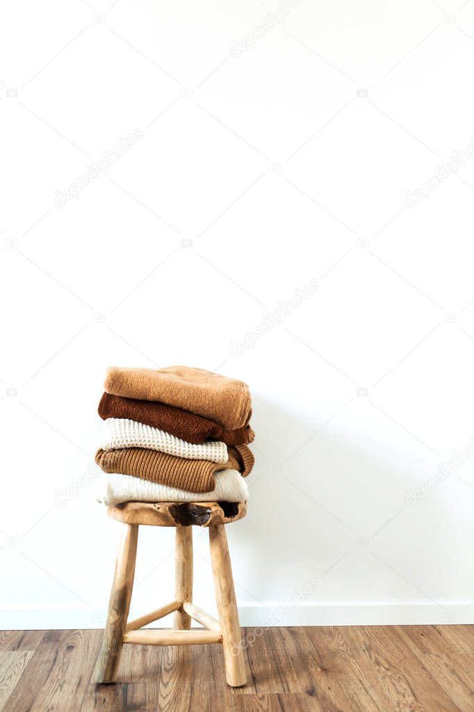 Warm winter women woolen knitted sweaters, pullovers stack on wooden stool at white background. Female modern clothes fashion blog concept.