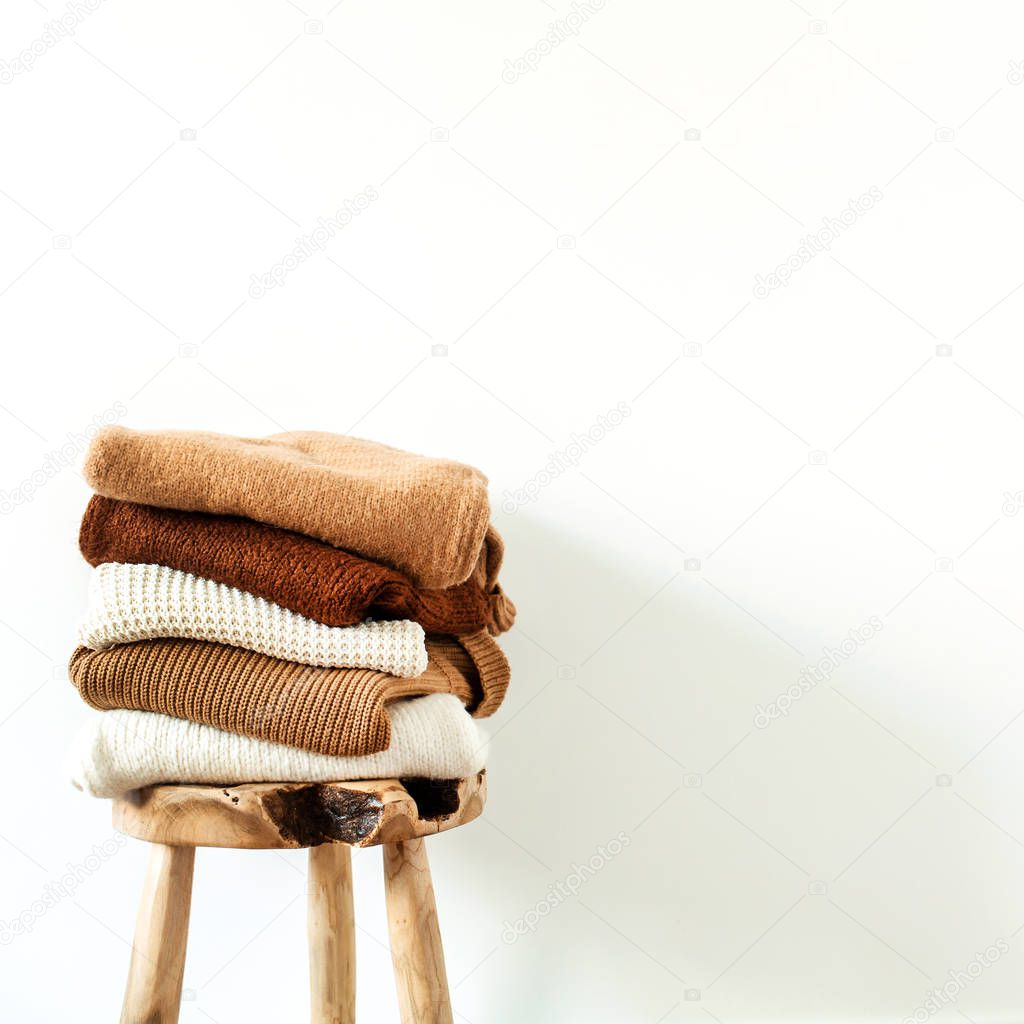 Women fashion composition. Warm winter female woolen knitted sweaters, pullovers stack on wooden stool at white background. Modern clothes concept for magazine, blog, social media.