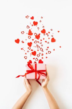 Beyaz arka planda pembe hediye kutusu ve kalp sembolü konfetisi tutan kadınların ellerinin olduğu Sevgililer Günü kompozisyonu. Düz konum, üst görünüm tatil konsepti.