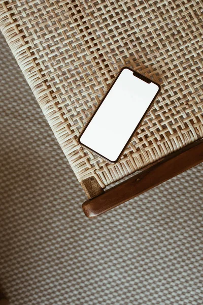 Blank screen phone on rattan wooden chair. Flat lay, top view copy space mockup template.