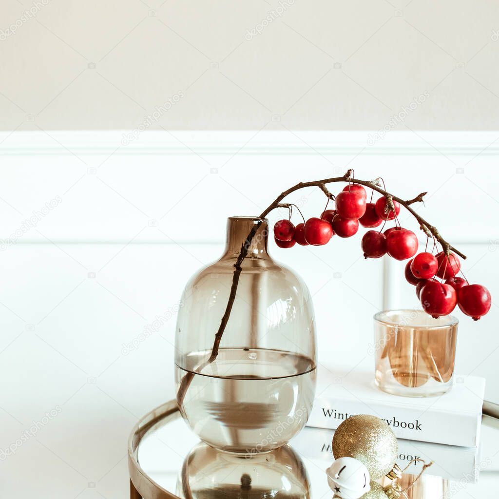 Modern classic interior design. Decorated living room. Bedside table with red berries bouquet in glass vase, book, candle in front of beige wall. Comfortable home apartment.