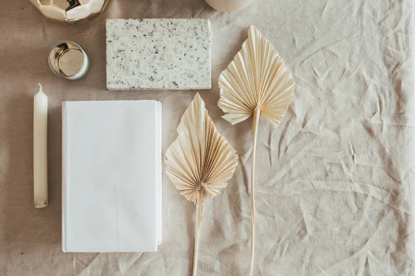 Lifestyle, still life composition with home decoration: book, fan leaves, granite brick, bust, candle, on beige linen table cloth. Flat lay, top view.