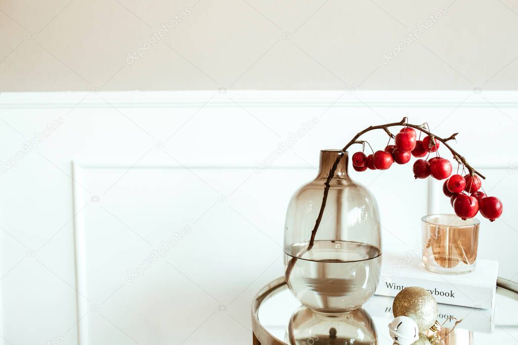 Modern classic interior design. Decorated living room. Bedside table with red berries bouquet in glass vase, book, candle in front of beige wall. Comfortable home apartment.