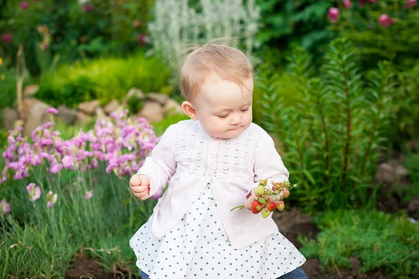Chica de 8 meses de edad bebé ucraniano europeo en un paseo por el jardín tiene una flor y fresas en sus manos —  Fotos de Stock