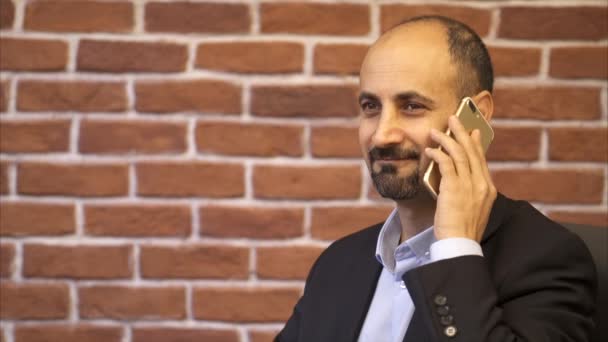Hombre de negocios con teléfono inteligente blanco habla y sonríe - Fondo de pared de ladrillo — Vídeos de Stock
