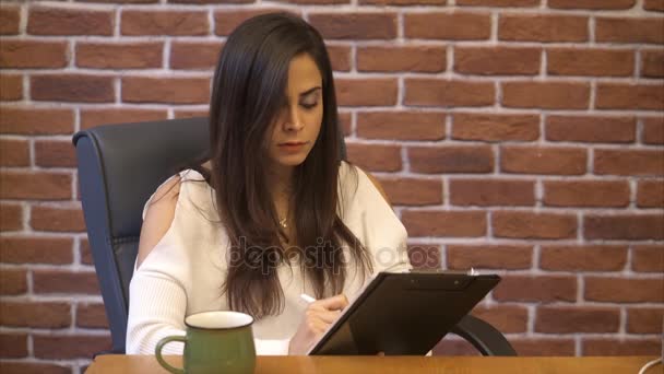 Geschäftsfrau notiert in einem Notizblock im Büro und trinkt Kaffee - Backsteinwand Hintergrund — Stockvideo