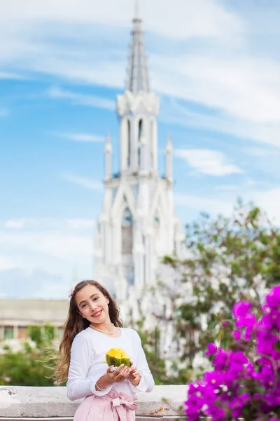 Hermosa joven en el puente de Ortiz comiendo un mango frente a la famosa iglesia gótica de La Ermita construida en 1602 en la ciudad de Cali en Colombia — Foto de Stock