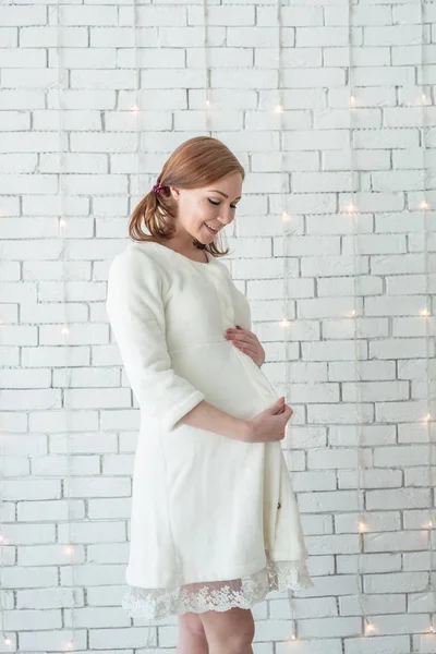 Jeune fille enceinte souriante et debout près du mur blanc — Photo