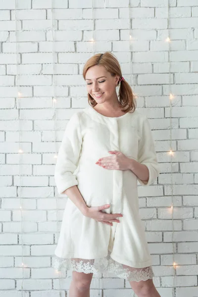 Jeune fille enceinte souriante et debout près du mur blanc — Photo
