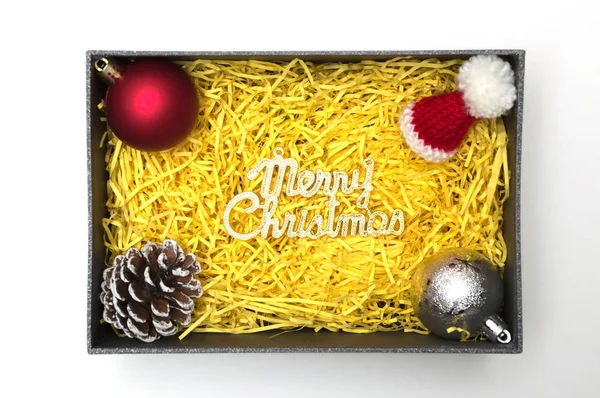 Merry christmas tekst en bont hoed, bal, dennenappel in zwarte geschenkdoos met gele verpakkingsmateriaal — Stockfoto