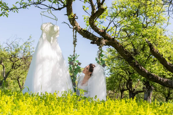 Een fantastische bruid in het park zit op de schommel en kijkt naar de trouwjurk — Stockfoto