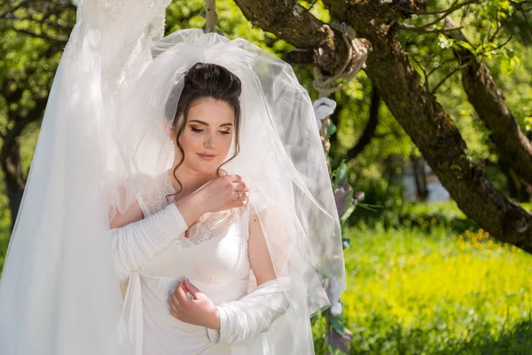 A fantastic bride in the park sits on the swing and looks at the wedding dress