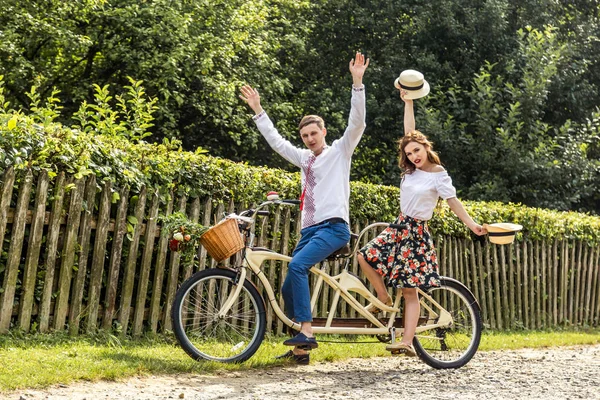 Bisiklet tandem Park ile genç çift. Gençler şapka almak onların yakışıklı ve gülümseme. Ağaç çitin arkasında