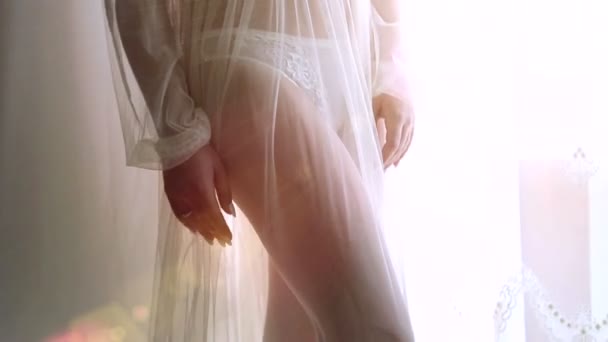 Una joven se para junto a la ventana con una camisa de noche. Piernas cerca de un hermoso modelo — Vídeos de Stock
