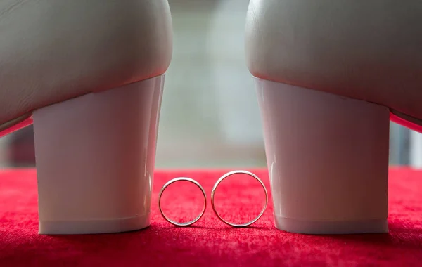 Dva snubní prsteny na pozadí červeného sametu. Nedaleké bílé svatební boty. — Stock fotografie