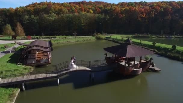 Vista aérea. Las novias felices están en un día soleado en un puente cerca del lago — Vídeos de Stock