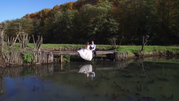 Las novias se sientan cerca del lago en un pequeño puente en el parque — Vídeo de stock