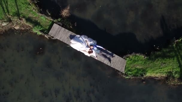 Bruiden liggen op een bruggetje in het park. Luchtfoto bovenop. Filmen een dron, vliegt de camera uit de buurt van de bruiden — Stockvideo