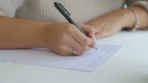 Mano femminile con una penna registra le informazioni su un pezzo bianco bianco di carta sdraiato sul tavolo . — Video Stock