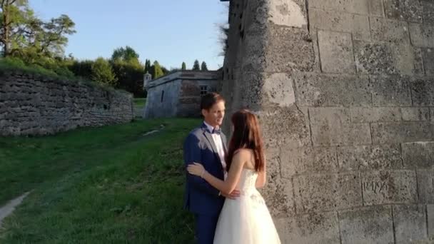 Felices recién casados al atardecer, de pie en la pared del castillo. El sol brilla en la cámara, los recién casados sonríen . — Vídeos de Stock