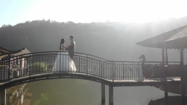 Jasne poranne słońce świeci nad nowożeńcami i przed kamerą. Nowożeńcy tańczą na moście nad jeziorem w pobliżu restauracji. Szczęśliwego słonecznego dnia ślubu. — Wideo stockowe