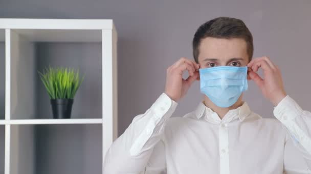 Jonge arts verwijdert een beschermend masker uit zijn gezicht in een gezellig kantoor en kijkt naar de camera. — Stockvideo