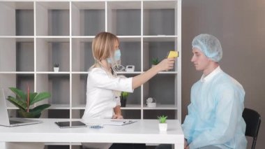 Beyaz önlüklü ve tıbbi maskeli bir doktor, tıbbi ofisteki bir hastanın ısısını kızıl ötesi temas olmayan termometreyle ölçer..