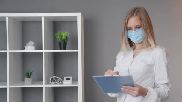 Pielęgniarka w białym płaszczu w masce medycznej, z niebieskim folderem i zapisem informacji w gabinecie medycznym — Wideo stockowe