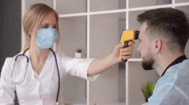 Beyaz önlüklü ve tıbbi maskeli bir doktor, tıbbi ofisteki bir hastanın ısısını kızıl ötesi temas olmayan termometreyle ölçer..