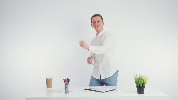 Danza de un empresario en el trabajo en el lugar de trabajo. Un joven empleado baila cerca del lugar de trabajo sobre un fondo blanco en una oficina luminosa — Vídeos de Stock