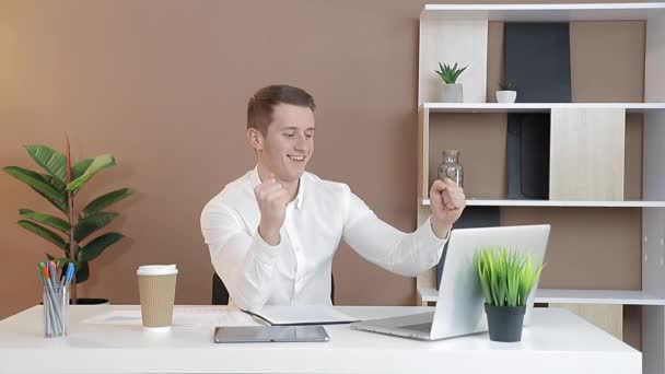 白いシャツを着たビジネスマンが職場に座ってコンピュータで働き誠実に笑っています. — ストック動画