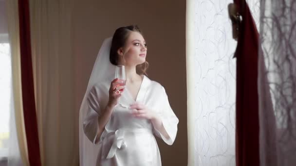 Ευτυχισμένη νύφη με ένα ποτήρι σαμπάνια στο παράθυρο την ημέρα του γάμου της. Ευτυχισμένα συναισθήματα μιας όμορφης νύφης με ρόμπα. — Αρχείο Βίντεο