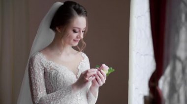 Mutlu gelin damat için bir düğün yaka çiçeğine bakar ve pencereye gülümser. Gelinlerin yüzüne sabah ışığı düşer..