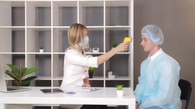 Beyaz önlüklü ve tıbbi maskeli bir doktor, tıbbi ofisteki bir hastanın ısısını kızıl ötesi temas olmayan termometreyle ölçer..