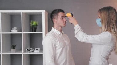 Beyaz önlüklü ve tıbbi maskeli bir doktor, tıbbi ofisteki bir hastanın ısısını kızıl ötesi temas olmayan termometreyle ölçer..