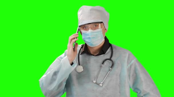 Молодой врач в медицинской маске и защитной медицинской одежде разговаривает по телефону. Снимок сделан на фоне Chrome Key. Chrome Kay высокое качество — стоковое видео