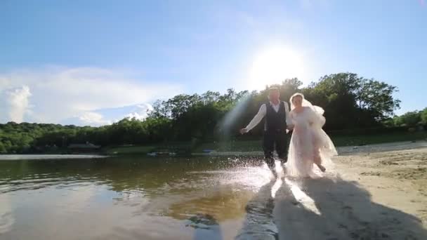 Friss házasok az esküvőjük napján, a vízen rohangálnak a tónál. Vízcseppek röpködnek. — Stock videók