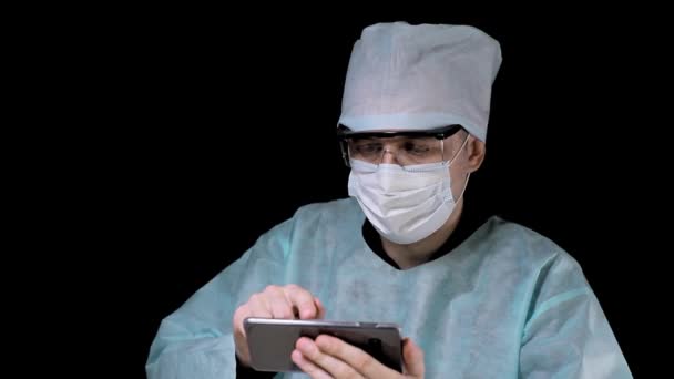Retrato de un médico cirujano que está sentado en una mesa y trabajando en un teléfono móvil sobre un fondo negro. Un médico que usa aparatos para trabajar . — Vídeos de Stock