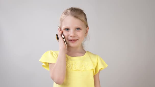 Una bambina è al telefono con un vestito giallo brillante su uno sfondo bianco. Conversazione emotiva al telefono di una bambina . — Video Stock