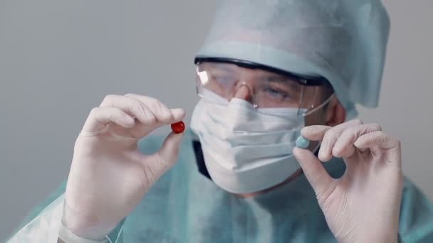 Médico vestido con una bata quirúrgica en una máscara médica con gafas apunta a dos tabletas de rojo y azul. Médico sosteniendo dos pastillas en guantes médicos blancos . — Vídeos de Stock